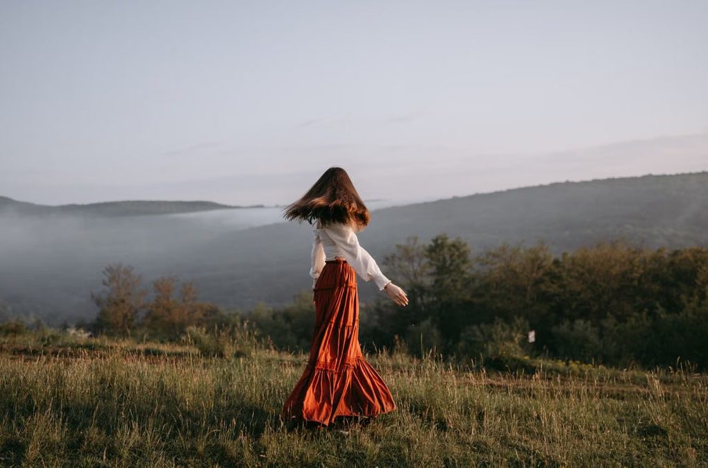 A woman in a flowing skirt walks through a misty meadow with hills in the background.