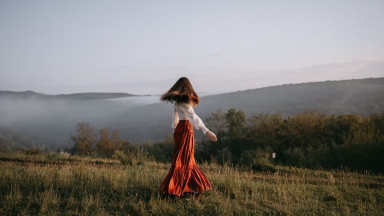 A woman in a flowing skirt walks through a misty meadow with hills in the background.